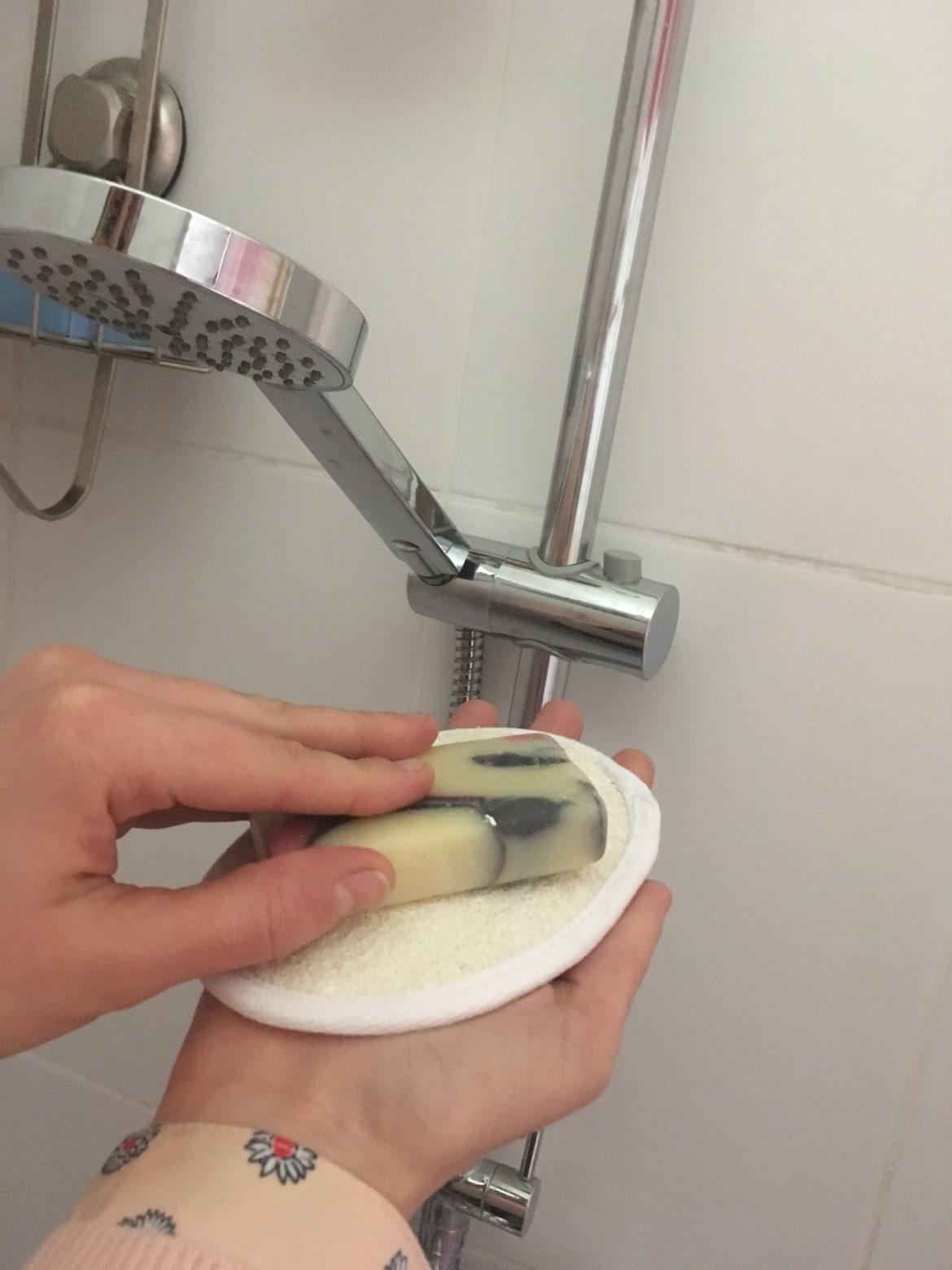 Une main dans un gant de loofah, sur lequel est posé un savon, devant un pommeau de douche