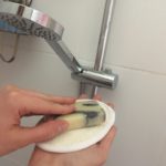 Une main dans un gant de loofah, sur lequel est posé un savon, devant un pommeau de douche