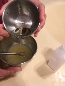 Une main verse de l'huile d'un bol dans un autre, contenant un gel couleur crème qu'une autre main est en train de fouetter