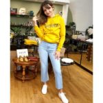 Ana est debout, en pied, vêtue d'un sweat jaune à capuche, d'un jean et de baskets blanches. Elle sourit et tire sur une de ses mèches de cheveux